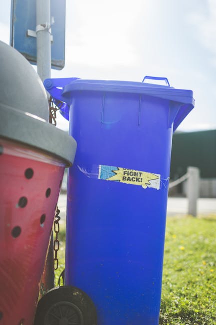 A bright blue wheeled rubbish bin with a closed lid, positioned on a grassy area outdoors, adjacent to a larger red bin with black dots on its side. The blue bin has a small rectangular sticker with t