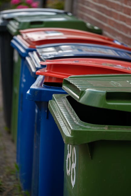 A bright blue wheeled rubbish bin with a closed lid, positioned on a grassy area outdoors, adjacent to a larger red bin with black dots on its side. The blue bin has a small rectangular sticker with t