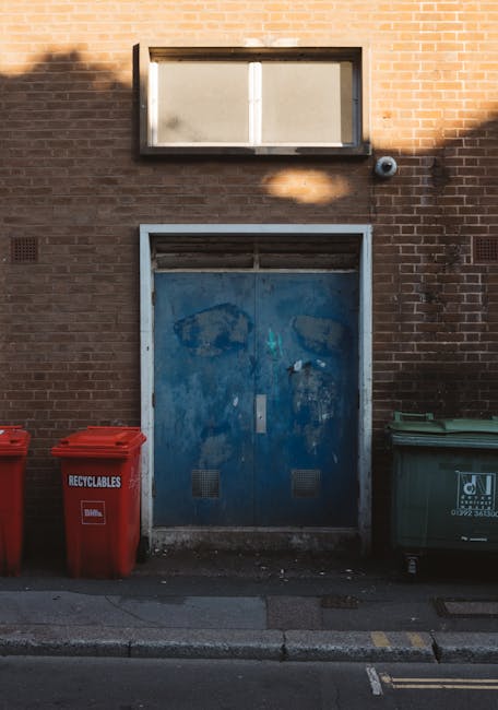 Two industrial skip containers are positioned outdoors against a background of a lightly overcast sky and a few leafless trees. The larger container on the left is painted in a dark blue hue with visi